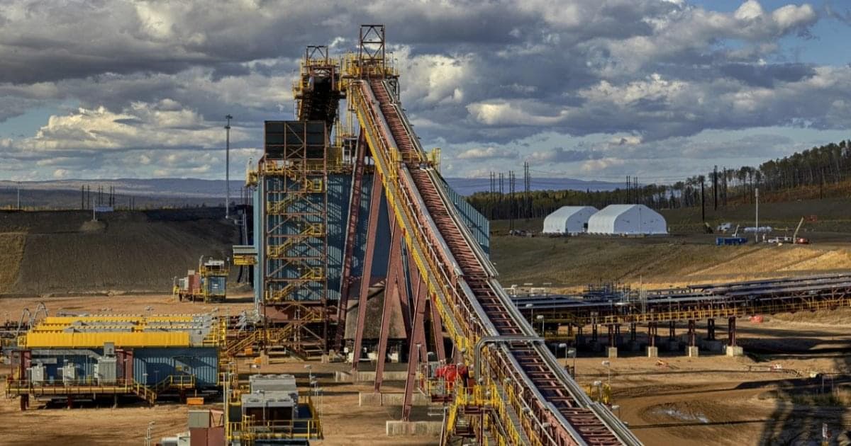 A large conveyor at an oil sands site. The conveyor belt is supported by large steel beams, and it curves upward. There are flat conveyor belts behind it, as well as buildings, grass and trees. The sky is cloudy and grey. 