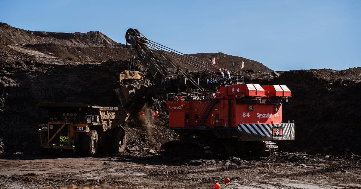 A large, red shovel at an oil sands mine. The shovel's bucket is loading a yellow haul truck with material from the mine.