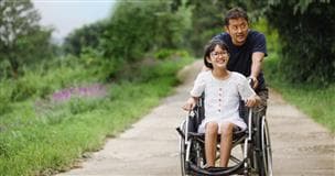 Un adulte qui pousse un enfant en chaise roulante sur un chemin. Le gazon est vert et des arbres bordent le chemin.