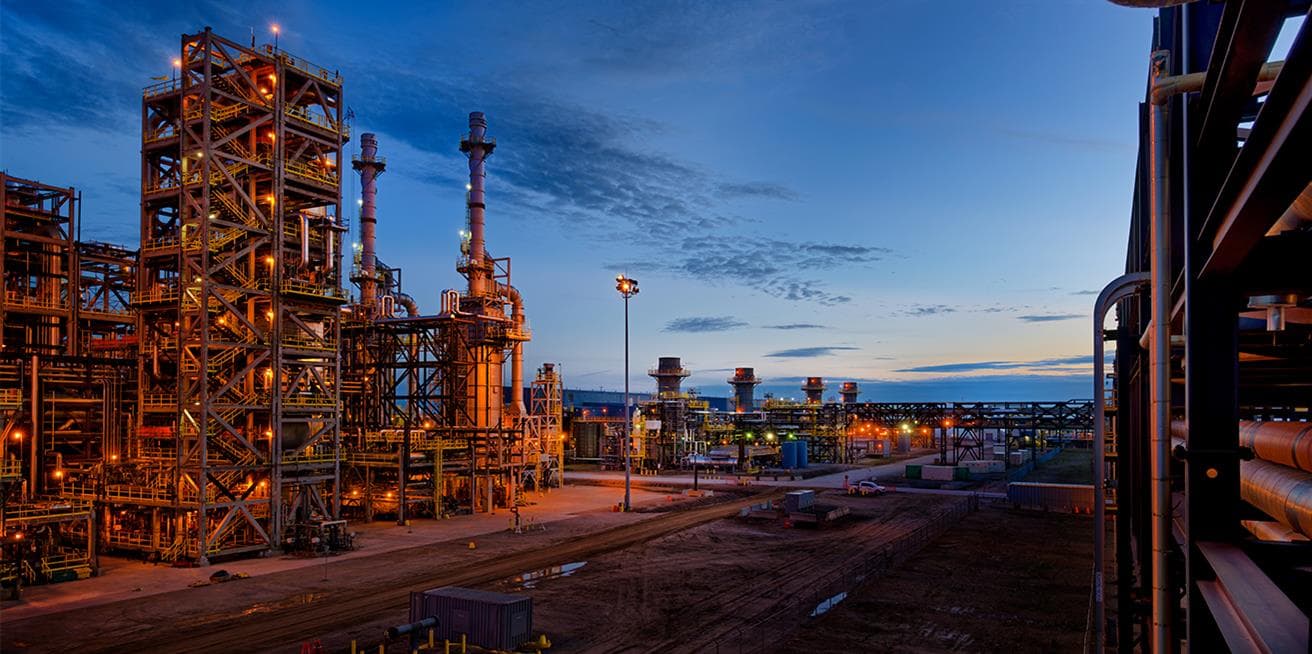 Suncor's Firebag in situ oil sands facility at dusk with illuminated processing units, pipelines and supporting infrastructure under an evening sky.