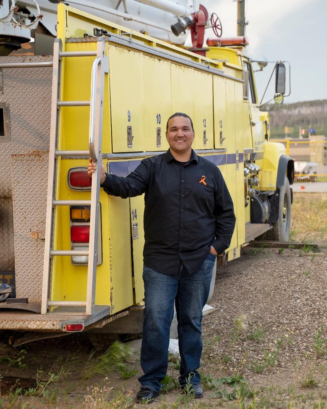 A man stands beside a large, yellow truck. He is holding on to the ladder attached to the truck and is wearing a black, long-sleeve shirt with an orange ribbon on it. 