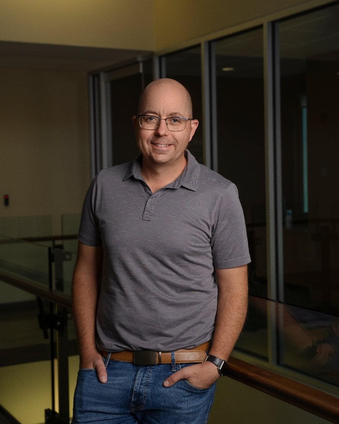 A man stands in a hallway inside an office building. He is wearing a grey t-shirt and jeans. 
