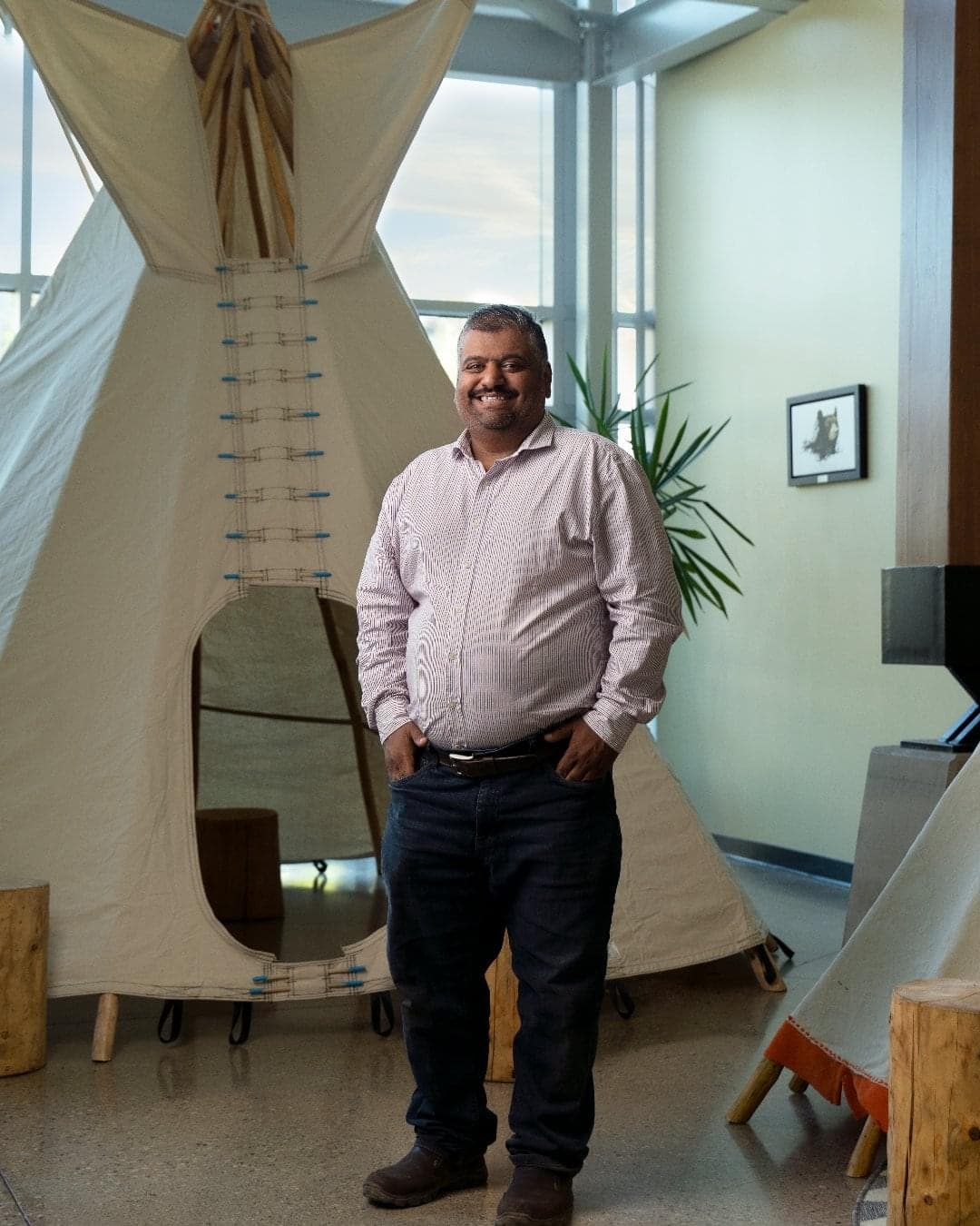 A man wearing a light-colour dress shirt and jeans stands in front of a tipi inside a building. 