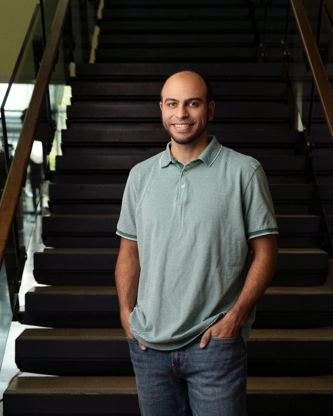 A man wearing a sage-colour t-shirt and jeans stands in front of a set of stairs.