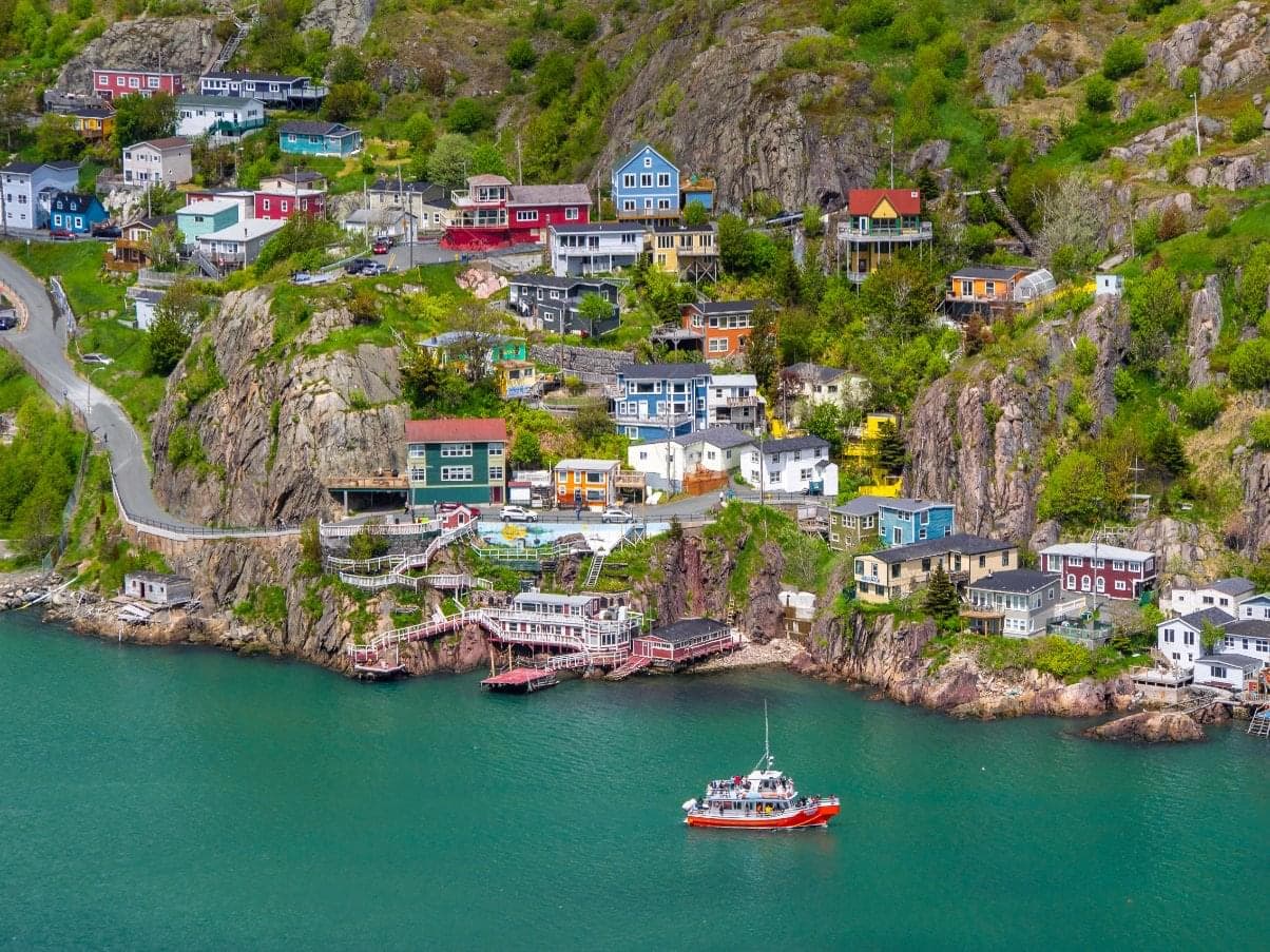 An aerial shot of St. John’s, N.L. harbour. There are colour houses along the rocky shore and a red boat in the water. The water is a dark green colour.