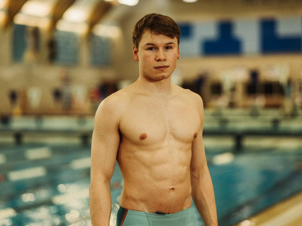 A young man standing by an indoor pool. He is swearing a light blue swimsuit and looking at the camera.