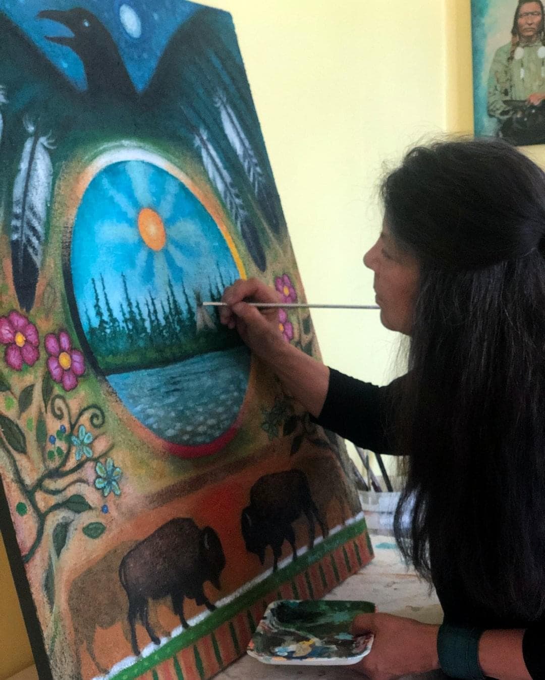 A woman with long, dark hair, paints a canvas that rests on an easel. The painting shows a black bird with its wings spread, a lake with trees a teepee and sun, wildflowers and two bison. 
