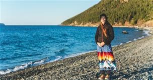 Kelly Young stands by the ocean. She is wearing a colourful, long ribbon skirt and a black jacket.
