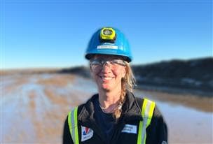 Sabrina Drake, debout sur une route minière à la mine Fort Hills de Suncor. Elle porte un manteau noir avec des bandes réfléchissantes et un casque bleu avec une lampe frontale. Il y a une route boueuse en arrière-plan.