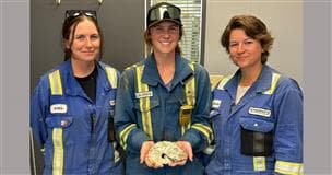 Trois femmes en combinaison sourient tandis que l'une tient un fossile d'ammonite.