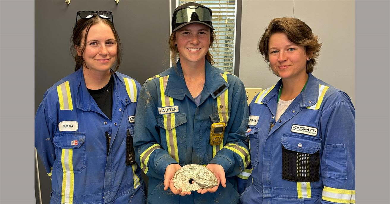 Trois femmes en combinaison sourient tandis que l'une tient un fossile d'ammonite.