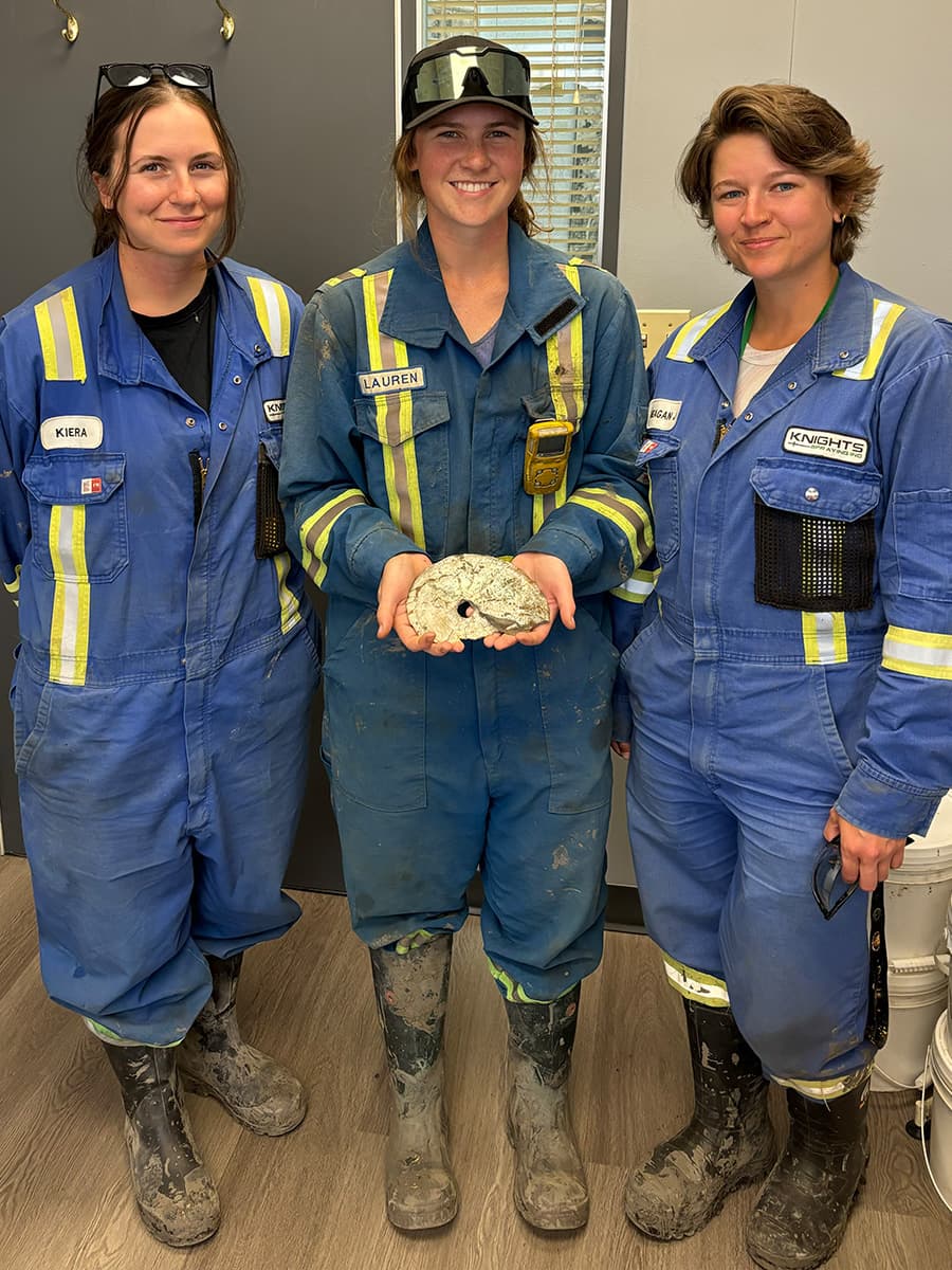 Trois femmes en combinaison sourient tandis que l'une tient un fossile d'ammonite.