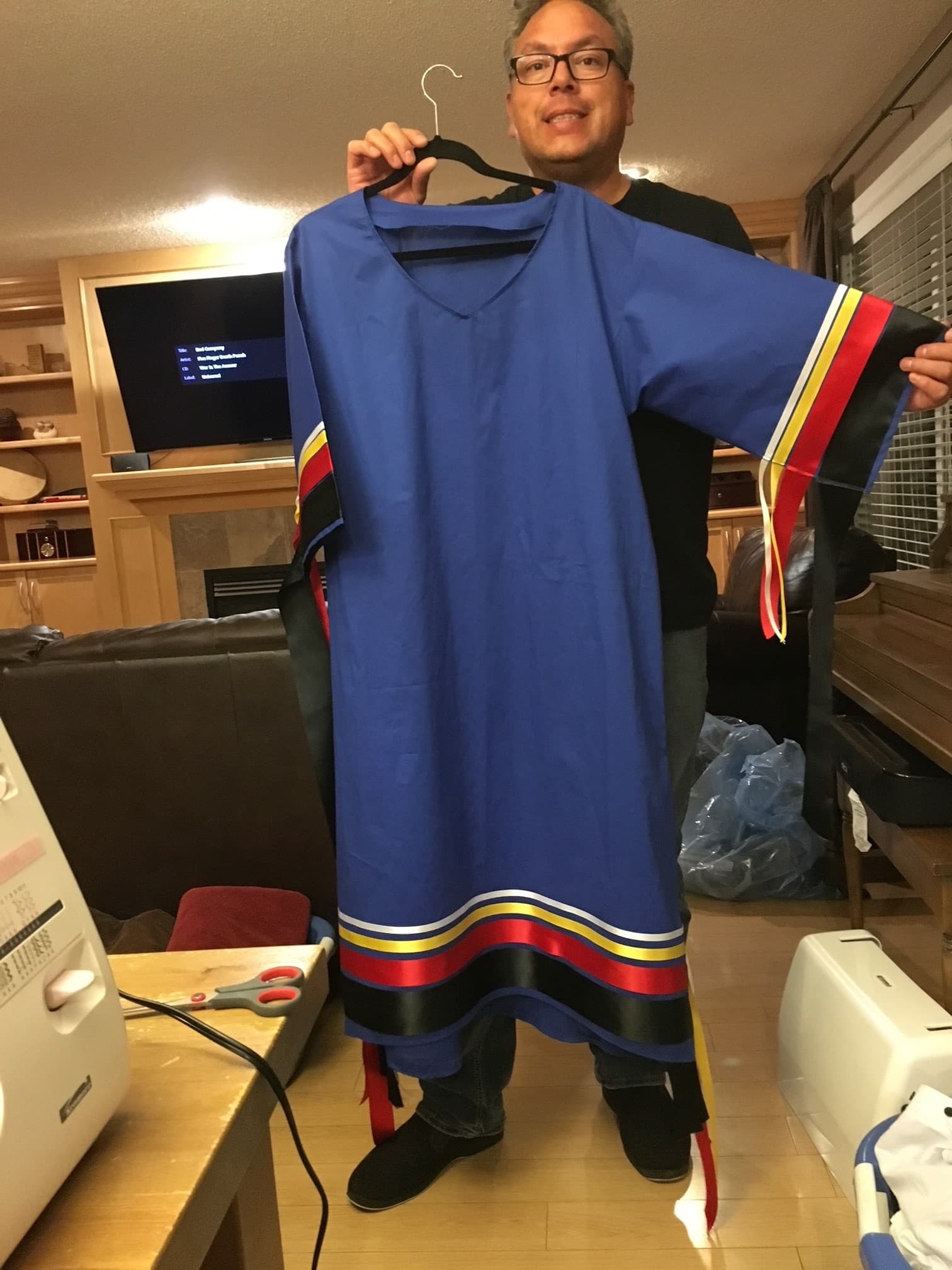 A man holding a blue dress with colourful ribbons sewn along the sleeves and bottom hem.