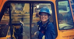 Une opératrice de grue assise dans le siège de l'opérateur d'une grue à tablier porteur Broderson de 9 tonnes. La grue est de couleur orange, et elle porte un ÉPI bleu, un casque bleu et des lunettes de sécurité.