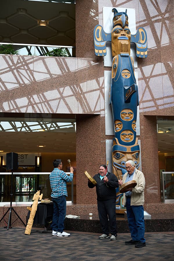 Klatle-bhi, Mike Lickers, and Elder Casey are singing and drumming in front of the totem pole