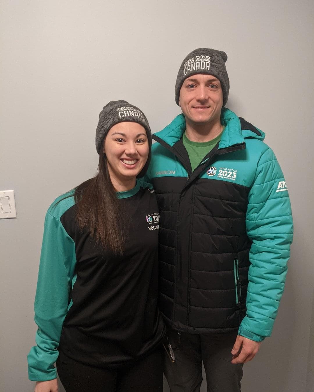 Deux personnes portant la tenue des Jeux d'hiver de l'Arctique de 2023 noire et turquoise se tiennent debout côte à côte. Souriant, ils portent tous les deux une tuque grise. 