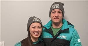 Two people wearing 2023 Arctic Winter Games apparel in black and turquoise, stand together. They are both wearing grey toques and smiling. 