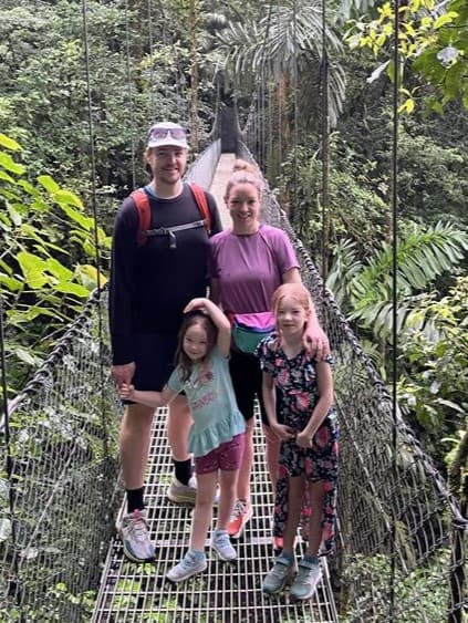 Julie with her husband and two daughters. They are standing on a rope bridge in the middle of a green forest.