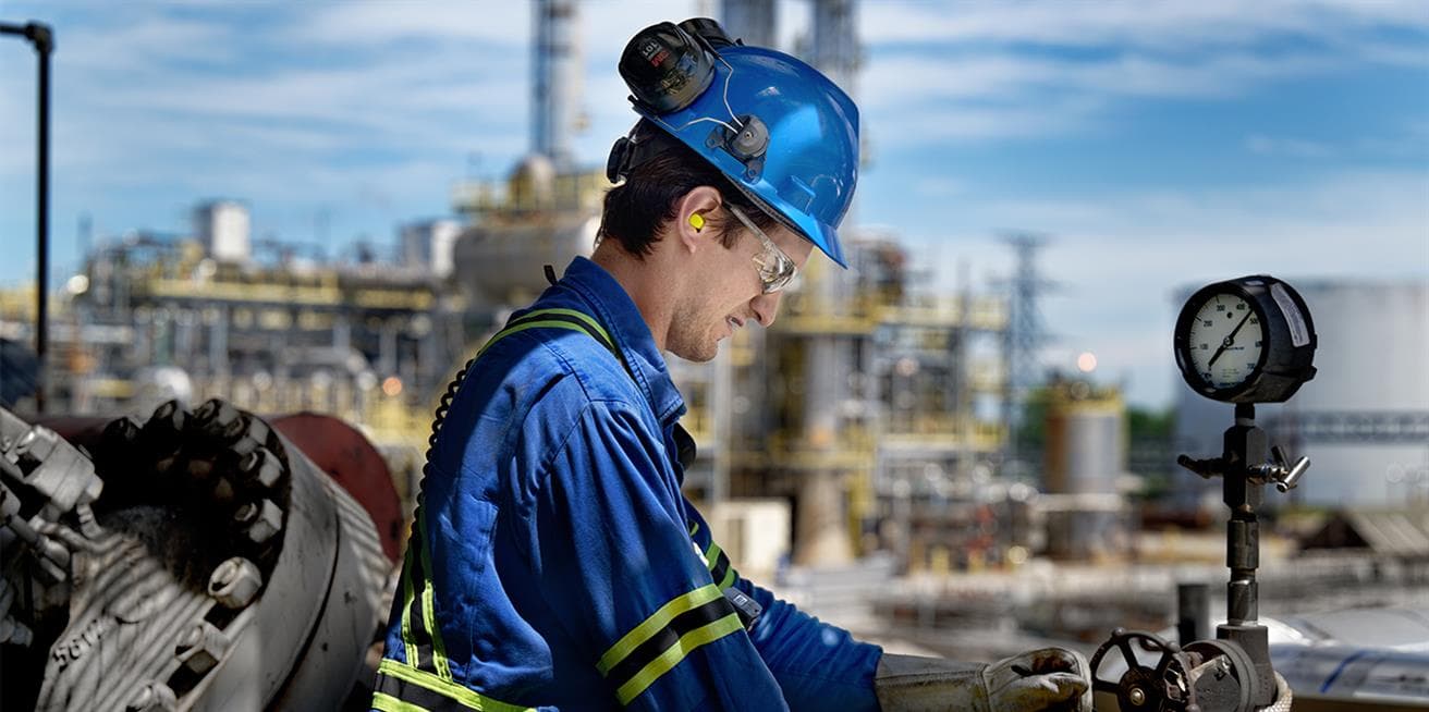 A worker at the Sarnia Refinery checks a yellow tag on a valve. He is wearing a blue hard hat, safety glasses, ear plugs, gloves and coveralls. Refinery operations and blue sky can be seen behind him in the background.