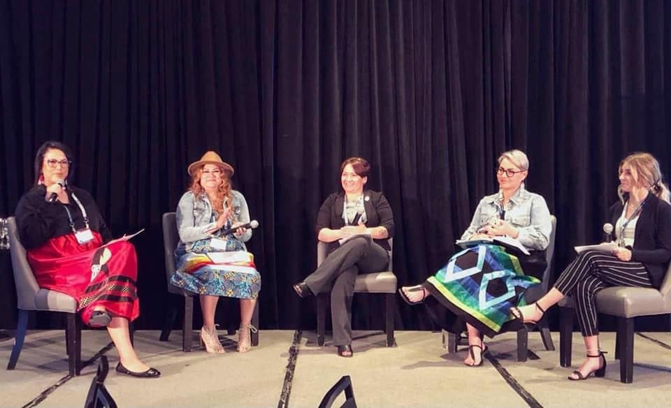 Cinq femmes sont assises sur une scène lors du panel du Forum d'apprentissage canadien. Lora porte une jupe autochtone traditionnelle vert et bleu et se trouve du côté droit de la scène.