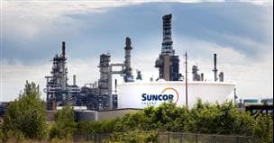 Aperçu de la raffinerie d'Edmonton en Alberta avec des tours en arrière-plan et un réservoir avec le logo de Suncor au premier plan, des arbres et un ciel nuageux.