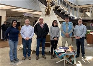 Un groupe de personnes debout devant un tipi à l'intérieur d'un bâtiment. Des plantes médicinales et objets traditionnels sont exposés.