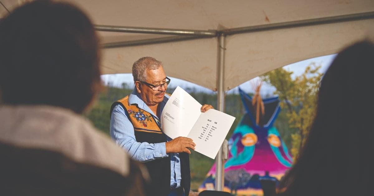 A man holding a book as he reads to a group of people under a large, white tent. There is a colourful tipi in the background.
