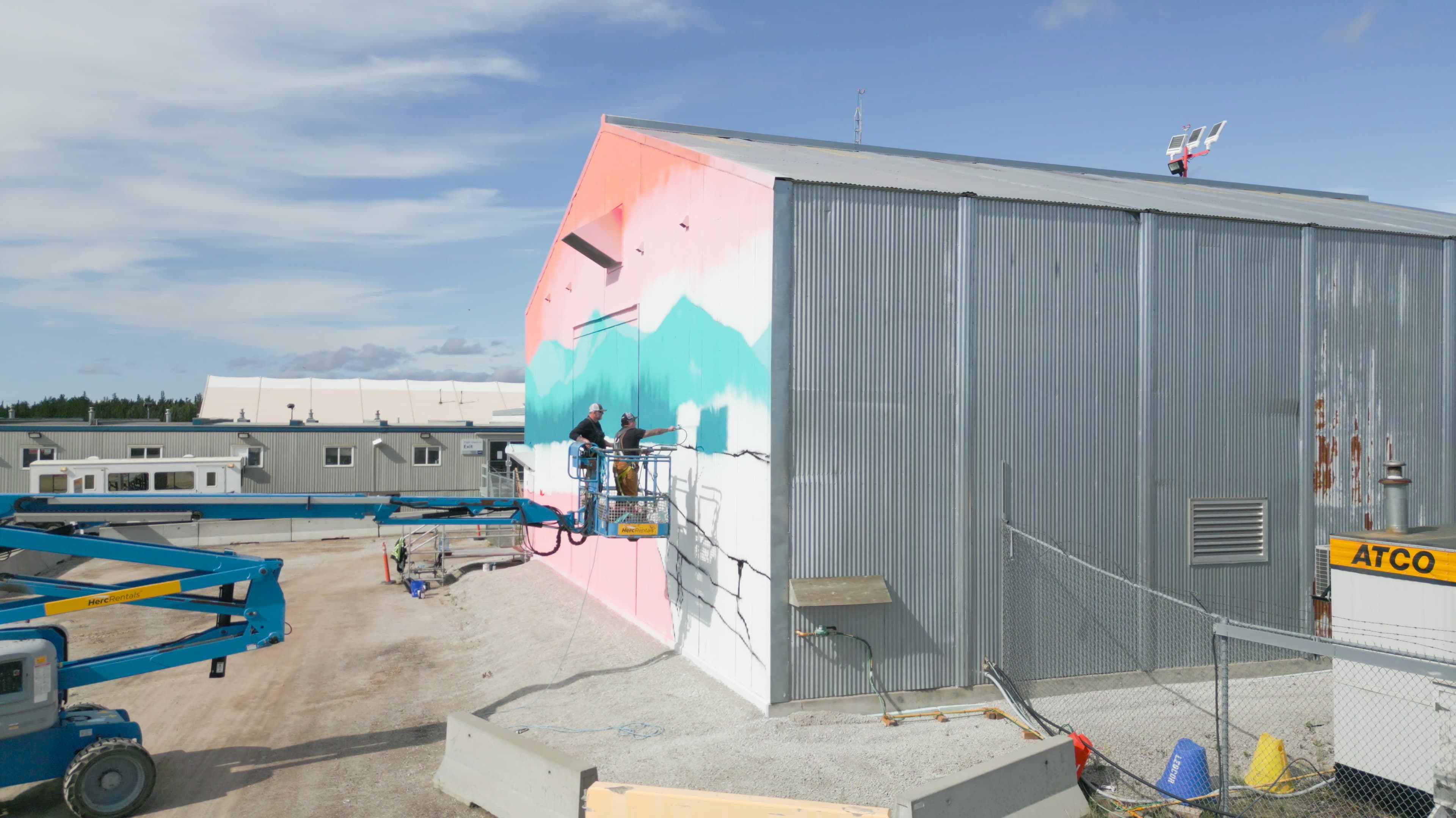 Keegan and another person are in a boom truck that is lifting them as Keegan paints the side of the firehall building. 