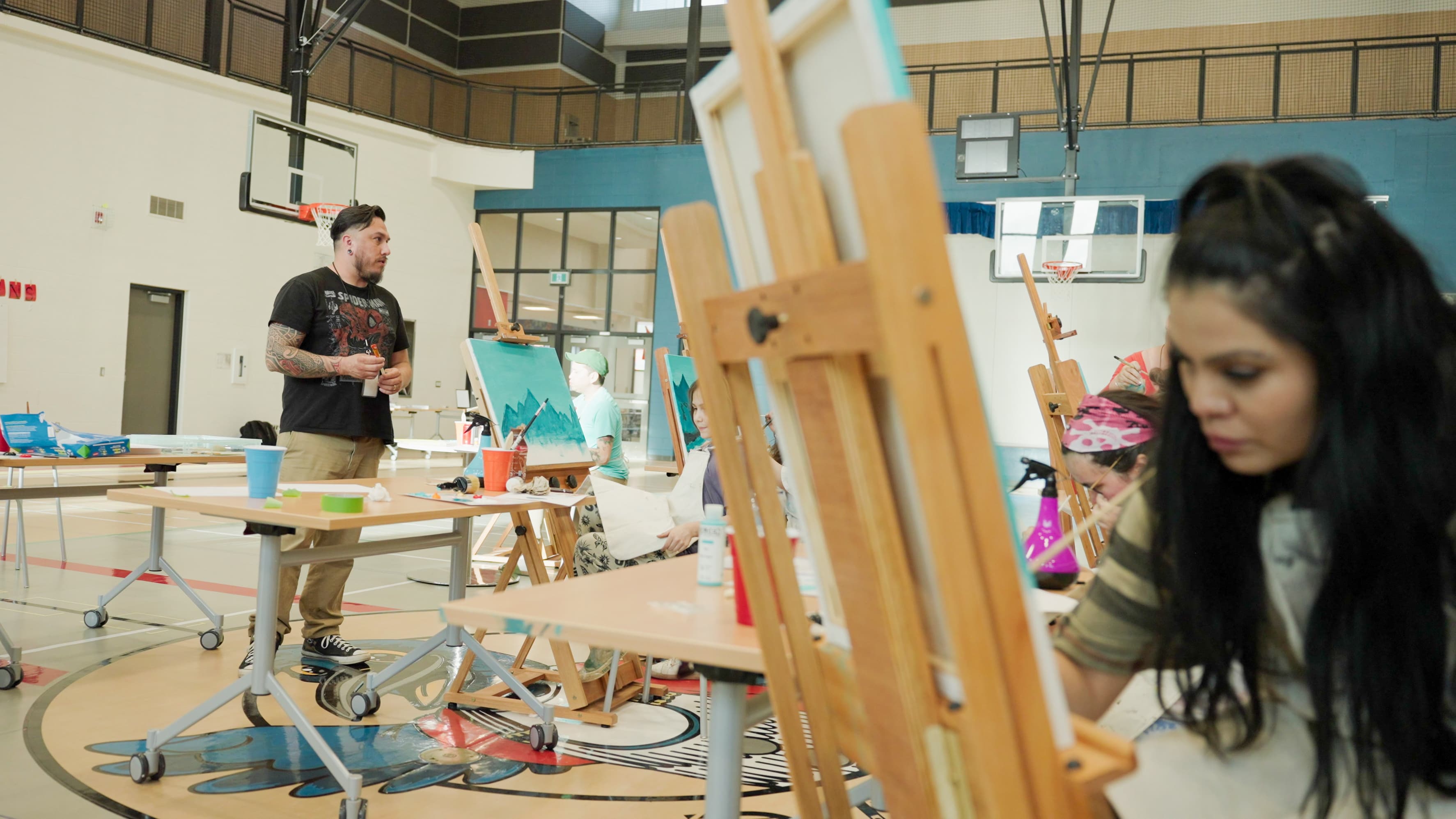 A group of people in a school gymnasium with easels and canvases. Keegan is standing at the front of the group.