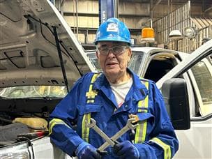 Un homme portant une combinaison bleue, des lunettes de sécurité et un casque bleu se penche sur un camion blanc. Le capot est ouvert et il tient des clés.
