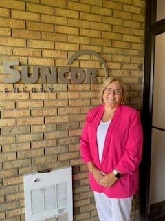 A woman wearing a pink blazer standing in front of a Suncor sign on a brick wall.