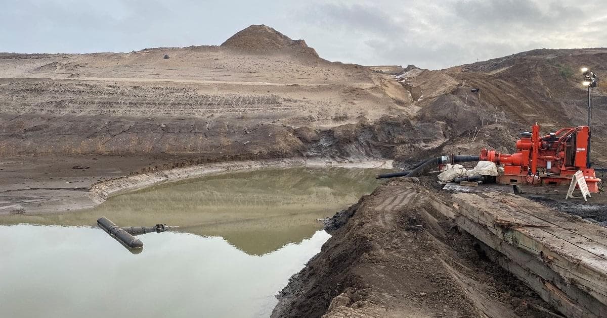 An after picture of a red pump on the land over a body of water. There are piles of sand and dikes around the water and hose line running from the pump to the water. The sky is grey with clouds. 