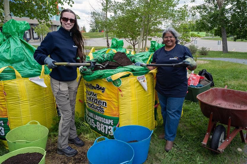 Two Suncor employees shovel dirt from large landscaping bags into wheelbarrows outside of a garden at the Alberta Children's Hospital in Calgary. They are wearing dark blue SunCares t-shirts and are smiling for the camera.