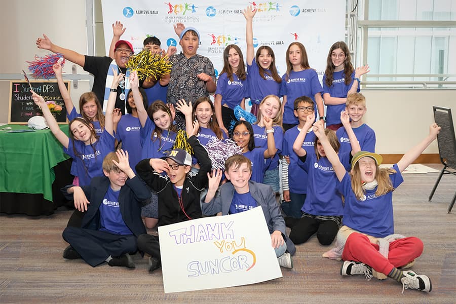A group of children in a classroom smiling and waving at the camera. They are all wearing matching purple shirts with the Achieve Gala logo embedded. 