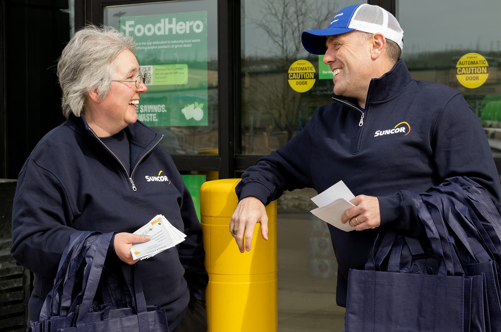 Deux employés de Suncor faisant du bénévolat à la banque alimentaire locale de Fort McMurray Wood Buffalo. Ils portent une veste bleu marine foncé de marque Suncor et sourient en se parlant. 