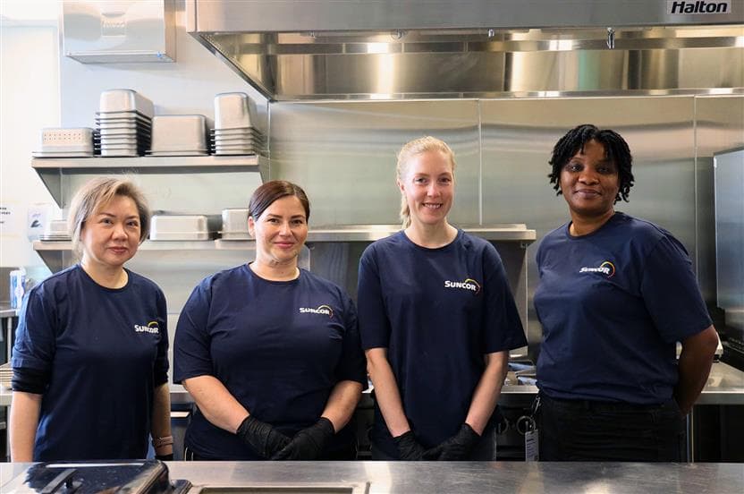 Quatre employées de Suncor debout derrière un comptoir en acier inoxydable dans une cuisine commerciale, portant des t-shirts Sun-Actif assortis et des gants sécuritaires pour les aliments.