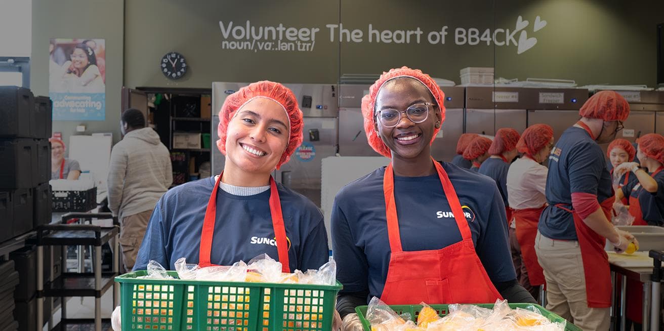 Suncor volunteers preparing and holding baskets of packaged food in a kitchen setting.