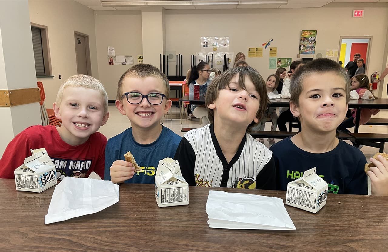 Four children sit at a cafeteria table with milk cartons and paper bags, eating snacks while other people sit at tables in the background.