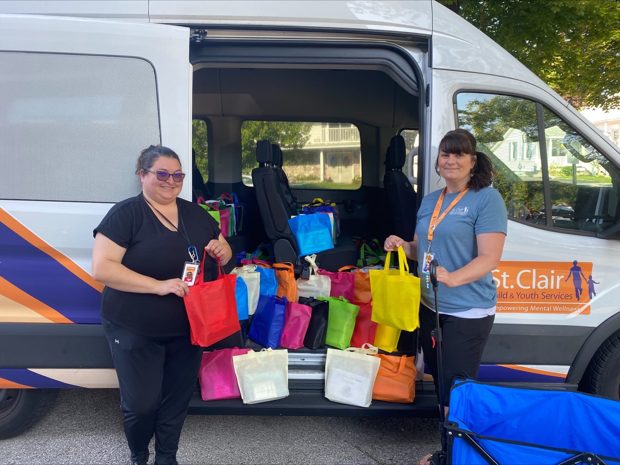 Two people stand beside an open van filled with colourful tote bags on a residential street, holding reusable bags in front of the vehicle. The van is decorated with graphics advertising St. Clair Child & Youth Services.