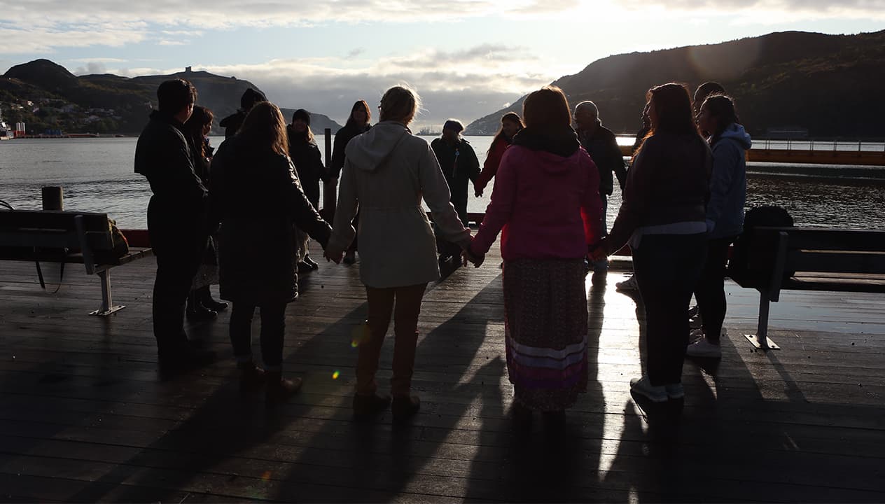 A group of people stand in a circle holding hands on a dock adjacent to a large body of water with mountains in the background. They are backlit by a setting sun. 