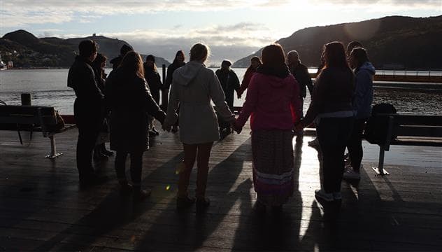 Un groupe de personnes forment un cercle en se tenant par les mains près d’un important cours d’eau avec des montagnes en arrière-plan. Ils sont éclairés à contre-jour par un soleil couchant.  