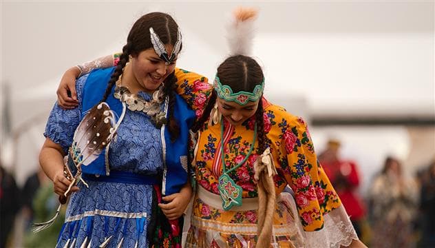 Deux femmes autochtones portant des vêtements autochtones colorés au 2025 Athabasca Tribal Council Festival. Elles dansent côte à côte et sourient.