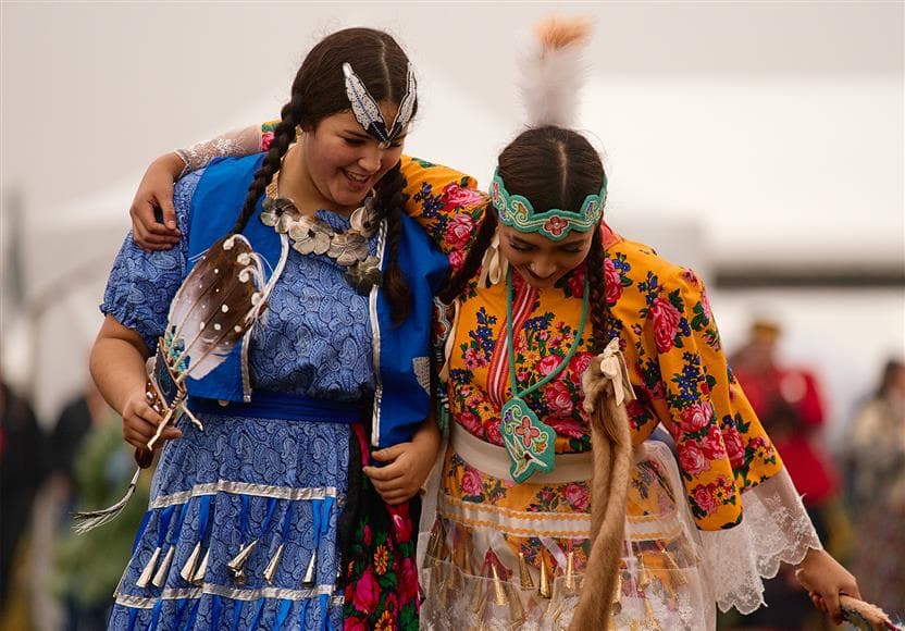 Two Indigenous women dressed in colorful Indigenous regalia at the 2025 Athabasca Tribal Council Festival. They are dancing side by side and smiling.