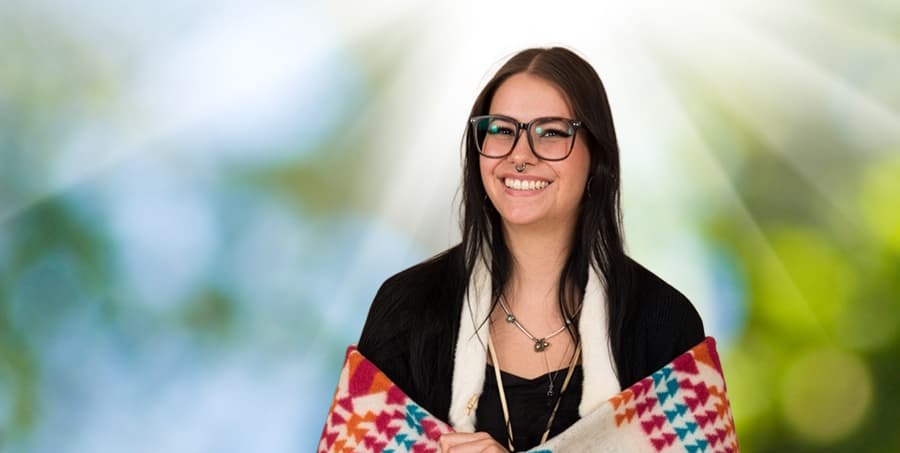 A headshot of a female Indigenous student. She is wrapped in a traditional blanket and is holding a feather. There is a vibrant array of colors in the background. 