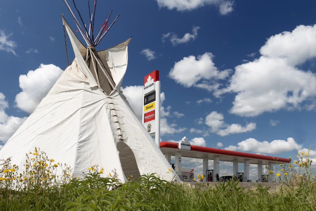 A teepee sits on a field of green grass in front of a Petro-Canada station. There are sunny blue skies in the background.