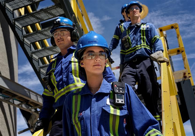 Four workers walking down a flight of stairs at the Sarnia Refinery. They are wearing blue hard hats, safety glasses, ear plugs and gloves.