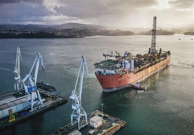 Une unité flottante de production, de stockage et de déchargement de couleur rouge fait son arrivée en cale sèche à Ferrol, en Espagne. Trois grues de couleur bleue se trouvent sur la tour du quai au-dessus du navire. Le soleil perce l'horizon nuageux où des montagnes et des zones urbaines se détachent derrière le navire.