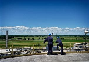 Deux travailleurs regardant vers une raffinerie. Ils portent une combinaison et de l’ÉPI, et il y a des champs verts et un ciel bleu derrière eux. 