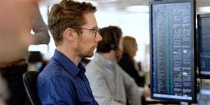 A male employee sitting at a desk working in Supply Trading. In the background there are computer monitors and other employees working.