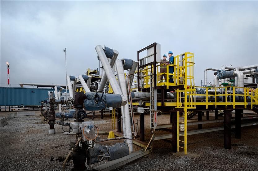 Two workers are standing on a platform by one of the wellheads at Suncor’s Firebag in situ site. They are wearing blue hard hats, ear plugs, safety glasses, coveralls and gloves.